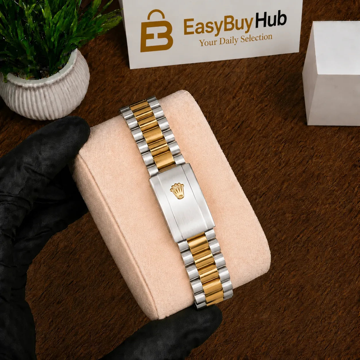 A two-tone metal watch band with a silver clasp featuring a crown emblem, resting on a beige display cushion. A hand wearing a black glove holds the cushion, with a wooden table, a small potted plant, and an EasyBuyHub sign in the background.