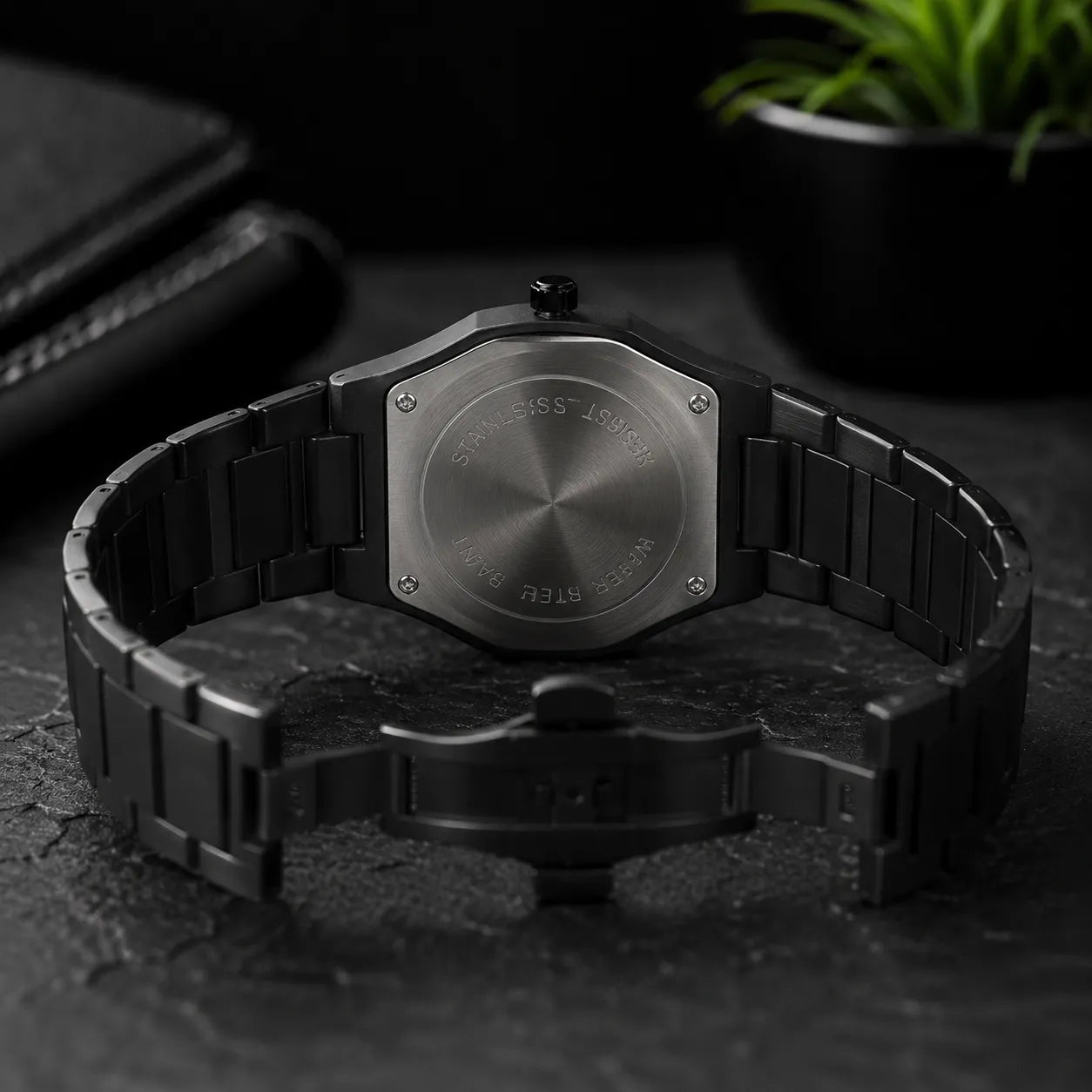 A black stainless steel watch with an integrated link bracelet, shown from the back on a dark textured surface with a blurred plant in the background.