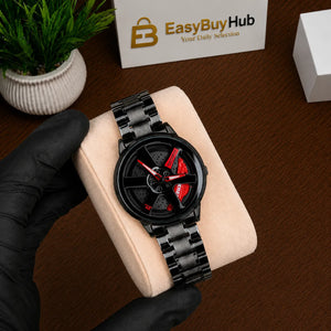 A black analog watch with a glossy black metal link bracelet and a red accent on the dial, held by a hand wearing a black glove on a beige cushion. The background shows a wooden table, a small potted plant, a branded EasyBuyHub box, and another white box.