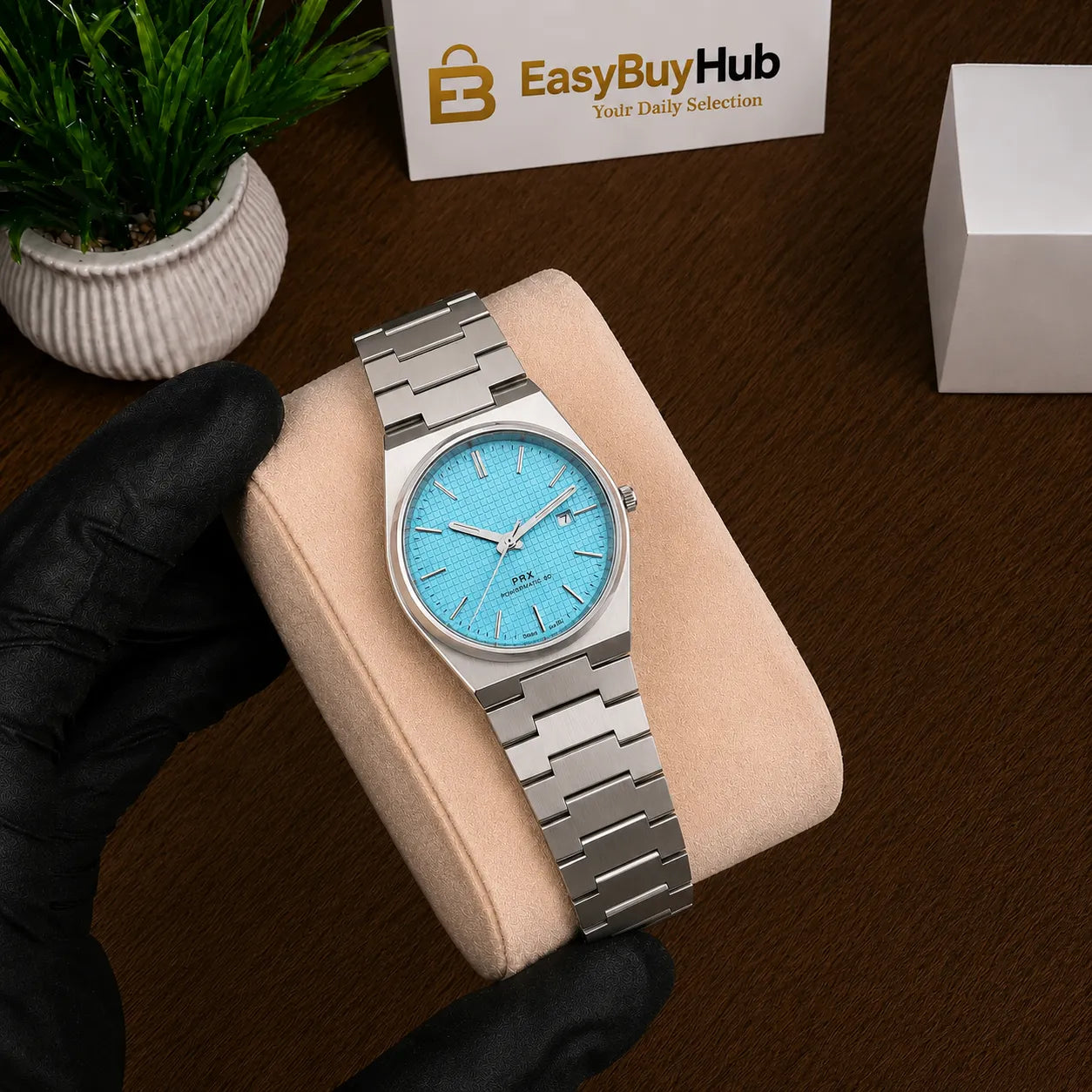 A silver stainless steel wristwatch with a light blue dial is displayed on a beige cushion held by a hand wearing a black glove. The background features a dark wood surface, a small potted plant, and a card for 'EasyBuyHub'.