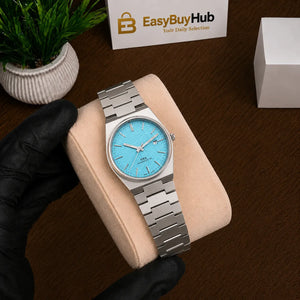A silver stainless steel wristwatch with a light blue dial is displayed on a beige cushion held by a hand wearing a black glove. The background features a dark wood surface, a small potted plant, and a card for 'EasyBuyHub'.