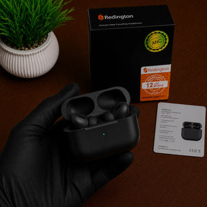 A hand wearing a black glove holds an open black Redington wireless earbud charging case, with the black product box, a small card, and a potted plant on a brown wooden surface.