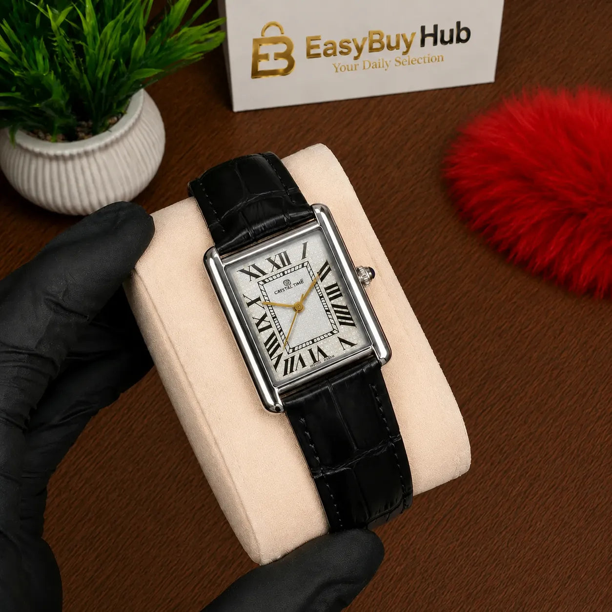 A rectangular analog watch with a black leather strap and silver-toned case, displayed on a cream cushion held by a gloved hand. The watch has a white dial with black Roman numerals.