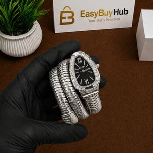 A silver-toned coiled bracelet watch with a black dial and crystal-embellished bezel, held in a black-gloved hand over a brown wooden surface. A small potted plant, a white display card reading EasyBuyHub, and a white box are visible in the background.