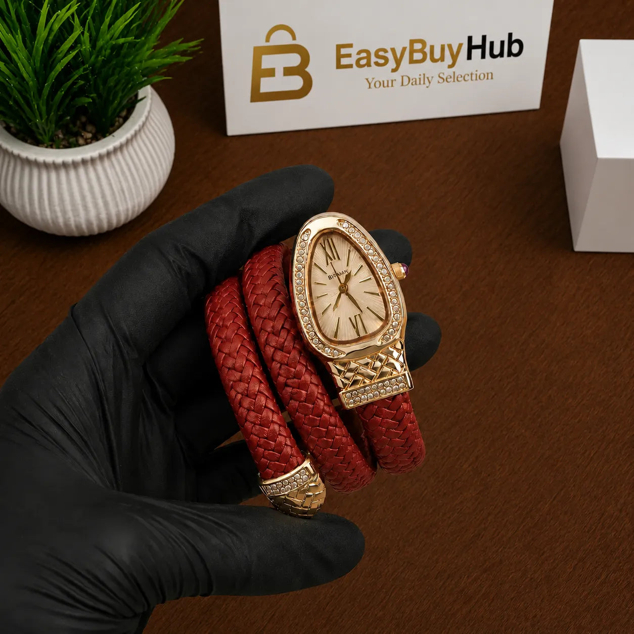 A red braided leather wrap watch with a gold-tone case and bezel, held in a hand wearing a black glove. The background shows a wooden table, a small potted plant, a branded EasyBuyHub card, and a white box.