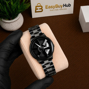 A black analog wristwatch with a matching black link bracelet is displayed on a beige cushion held by a black-gloved hand. The watch has a round case with a skeleton-style dial showing white hands and inner details.