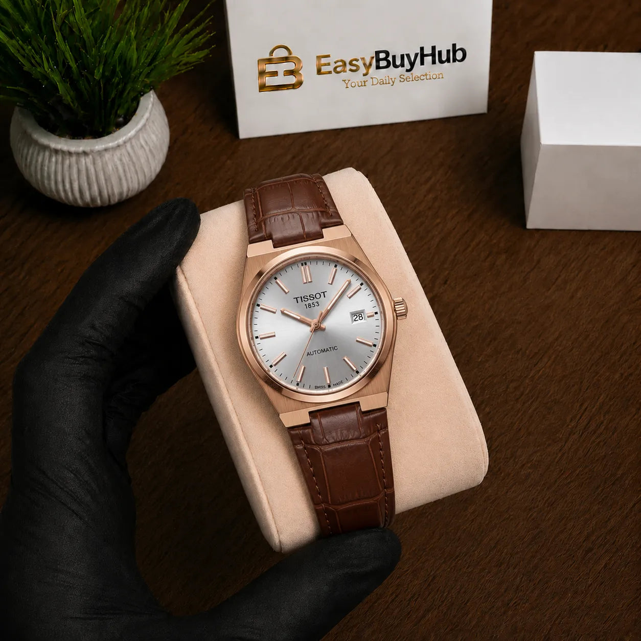A wristwatch with a rose gold case and brown leather strap displayed on a beige cushion, held by a hand wearing a black glove. The background shows a wooden table, a small potted plant, and a box labeled EasyBuyHub.