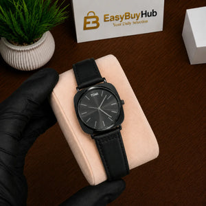 A black Tomsi watch with a black leather strap and a dark gray dial, resting on a light pink watch cushion held by a hand in a black glove. The background shows a dark wooden table, a small potted plant, and a box with the 'EasyBuyHub' logo.