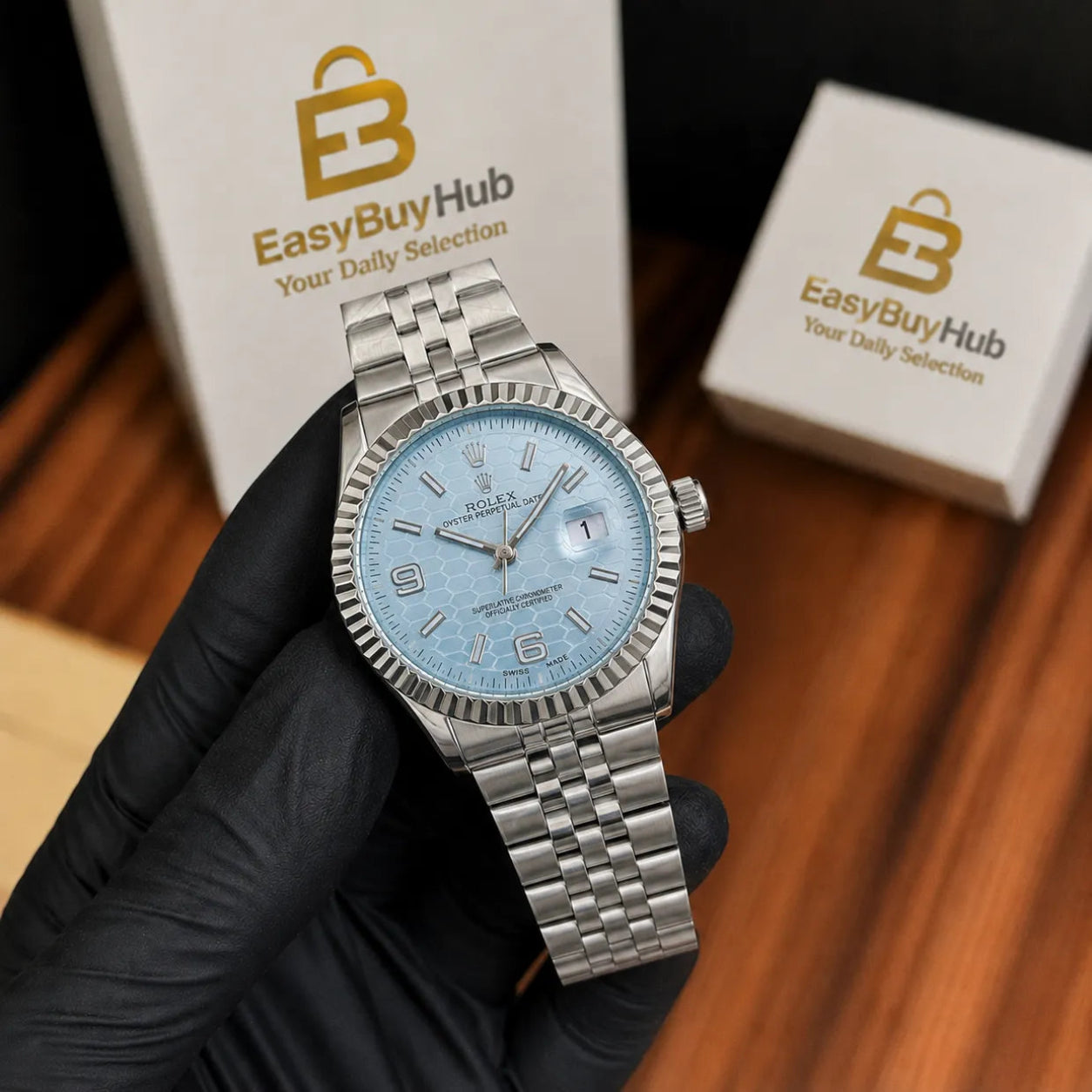 A Rolex Datejust watch with a light blue dial and silver stainless steel Oyster bracelet, held by a hand wearing a black glove. Two white EasyBuyHub boxes are visible in the background on a wooden surface.