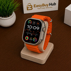 An orange silicone watch band mounted on a silver smartwatch, displayed on a beige stand on a dark brown wooden table. In the background, there is a small potted plant and a sign that reads 'EasyBuy Hub'.