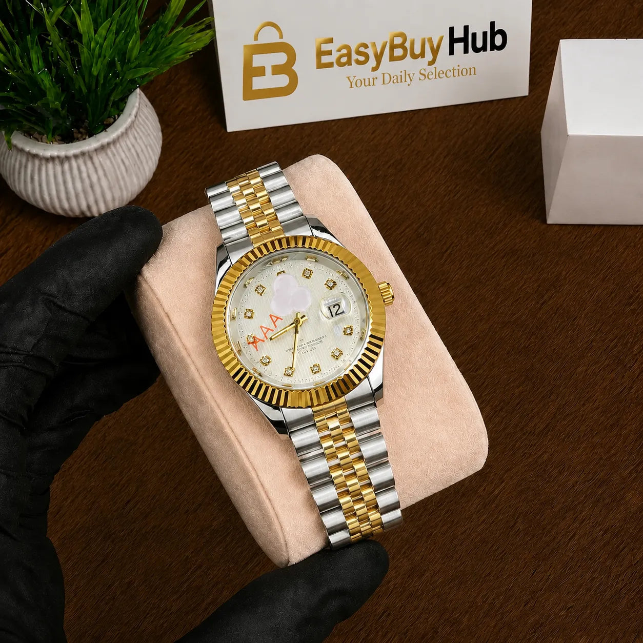 A gold and silver stainless steel watch with a white dial and crystal rhinestone hour markers, held by a hand wearing a black glove on a beige cushion. The background shows a wooden table, a small potted plant, and a card with the 'EasyBuyHub' logo.