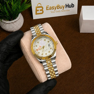 A gold and silver stainless steel watch with a white dial and crystal rhinestone hour markers, held by a hand wearing a black glove on a beige cushion. The background shows a wooden table, a small potted plant, and a card with the 'EasyBuyHub' logo.