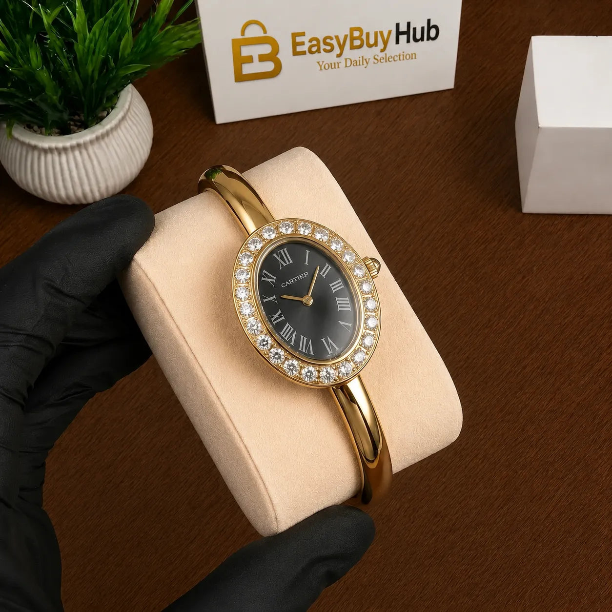 A gold bangle watch with a black dial and crystal-embellished bezel, held on a beige display cushion by a gloved hand. A branded EasyBuyHub box and a small potted plant are visible in the background on a brown tabletop.