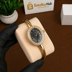 A gold bangle watch with a black dial and crystal-embellished bezel, held on a beige display cushion by a gloved hand. A branded EasyBuyHub box and a small potted plant are visible in the background on a brown tabletop.