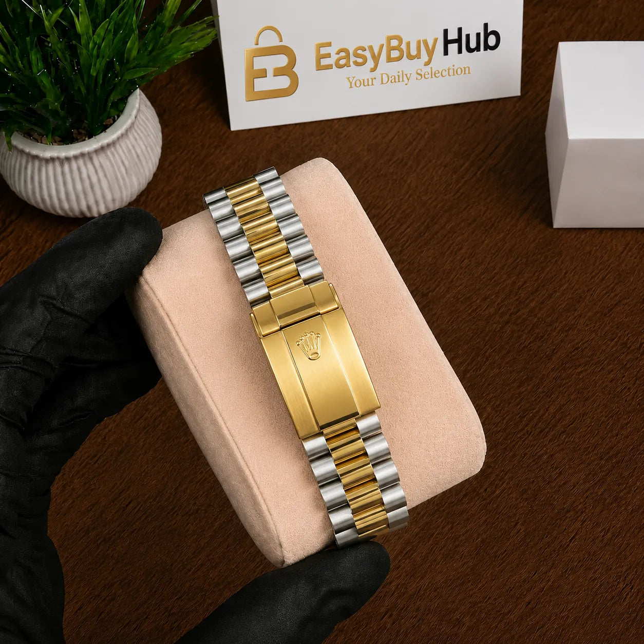 A two-tone gold and silver metal bracelet watch band is displayed on a beige velvet jewelry cushion, held by a hand wearing a black glove. The background shows a wooden table, a small potted plant, and a branded EasyBuyHub card.