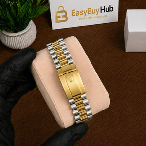 A two-tone gold and silver metal bracelet watch band is displayed on a beige velvet jewelry cushion, held by a hand wearing a black glove. The background shows a wooden table, a small potted plant, and a branded EasyBuyHub card.