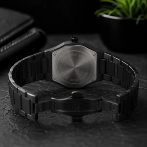 A black stainless steel watch with an integrated link bracelet, shown from the back on a dark textured surface with a blurred plant in the background.
