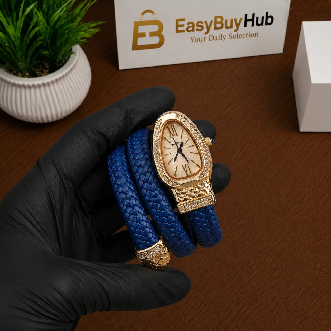 A gold-tone watch with an oval crystal case and a blue braided leather strap, held in a black-gloved hand over a brown wooden surface. A small potted green plant and a white sign reading EasyBuyHub are visible in the background.