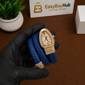 A gold-tone watch with an oval crystal case and a blue braided leather strap, held in a black-gloved hand over a brown wooden surface. A small potted green plant and a white sign reading EasyBuyHub are visible in the background.