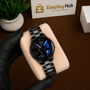 A black analog watch with a matching black link bracelet and a blue accent on the dial, displayed on a beige cushion held by a hand wearing a black glove. The background shows a wooden table, a small potted plant, and an EasyBuyHub box.