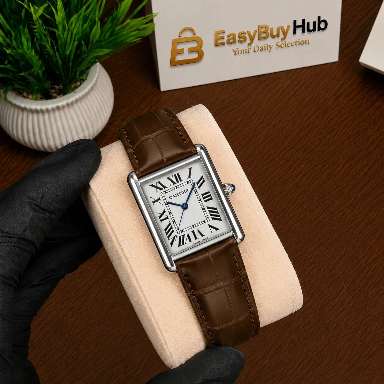 A Cartier Tank watch with a brown leather strap and silver case, held by a hand wearing a black glove on a beige display cushion. A small potted plant and a 'EasyBuyHub' card are visible in the background on a wooden table.