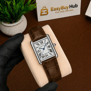A Cartier Tank watch with a brown leather strap and silver case, held by a hand wearing a black glove on a beige display cushion. A small potted plant and a 'EasyBuyHub' card are visible in the background on a wooden table.