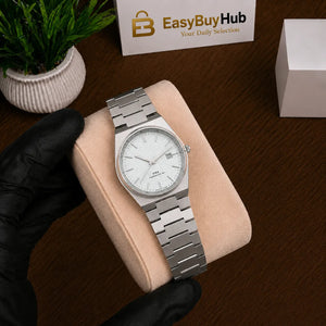 A silver stainless steel analog watch with a white dial and date window, resting on a beige cushion held by a hand wearing a black glove. A wooden table, a small potted plant, and an EasyBuyHub box are visible in the background.