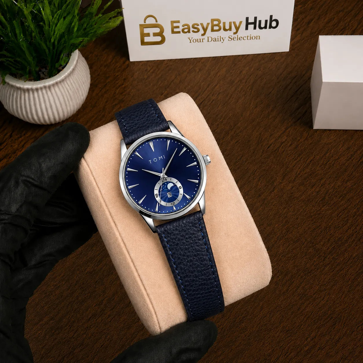 A wristwatch with a blue textured strap and a blue dial, displayed on a beige cushion. A hand wearing a black glove holds the cushion.