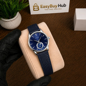 A wristwatch with a blue textured strap and a blue dial, displayed on a beige cushion. A hand wearing a black glove holds the cushion.