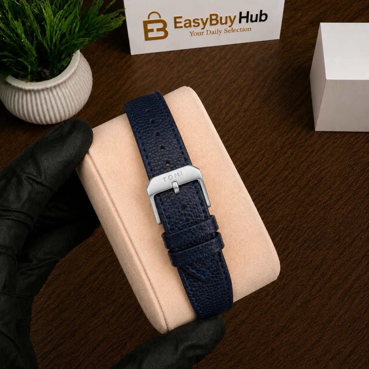 A navy blue textured leather watch strap with a silver buckle, displayed on a beige watch cushion held by a hand in a black glove, on a dark wooden table.