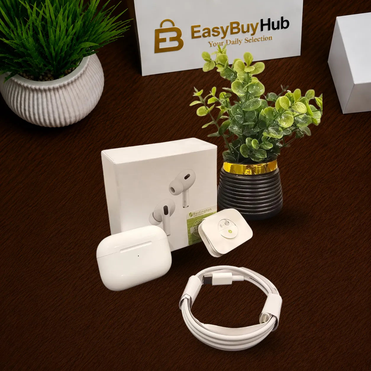 A wireless earbud charging case set with its white retail box, a white charging cable, a small white square accessory, and a small round tracker-like device, arranged on a dark brown tabletop with decorative plants and a branded EasyBuyHub sign in the background.
