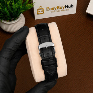 A black crocodile-embossed leather watch strap with a silver-toned metal buckle, displayed on a beige watch cushion held by a hand wearing a black glove, set on a wooden table.