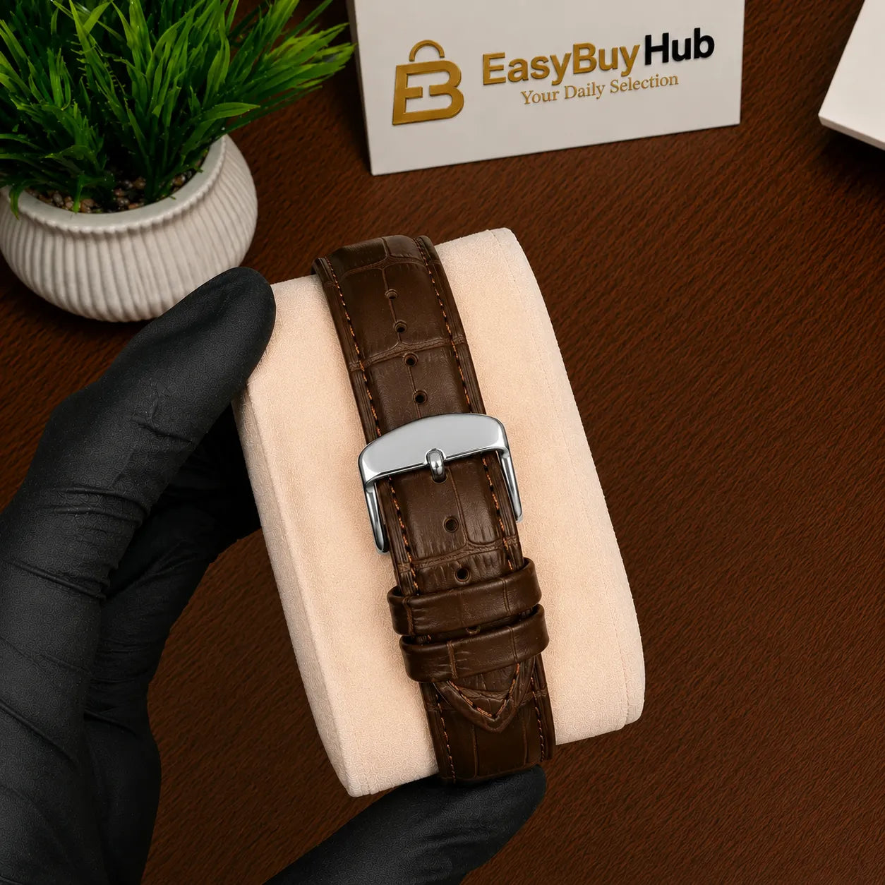 A brown crocodile-embossed leather watch strap with a silver buckle, held by a hand wearing a black glove on a beige display cushion over a wooden desk. A small potted plant and a card reading 'EasyBuyHub' are visible in the background.