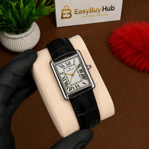 A rectangular analog watch with a black leather strap and silver-toned case, displayed on a cream cushion held by a gloved hand. The watch has a white dial with black Roman numerals.