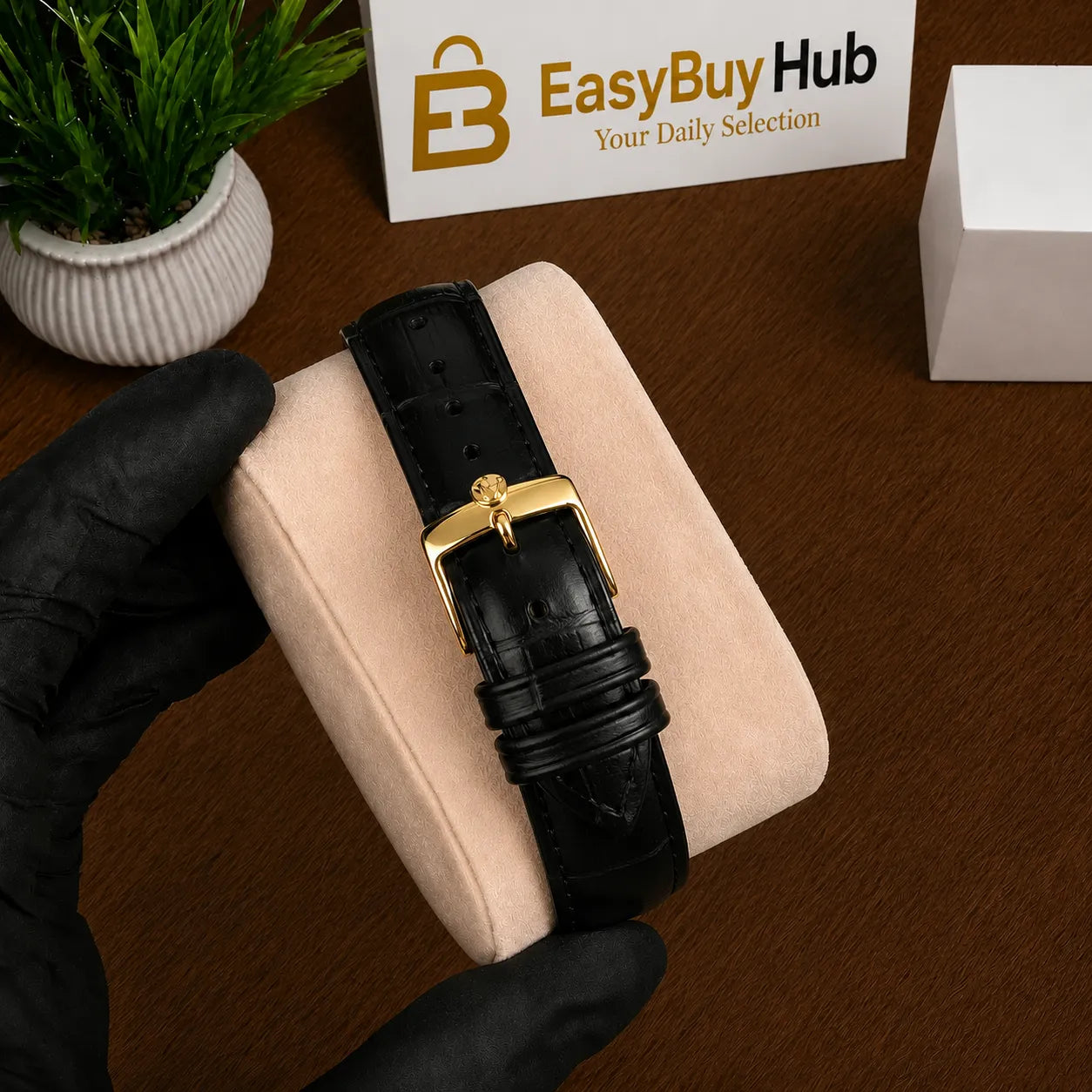 A black alligator leather watch strap with a gold buckle, held by a hand wearing a black glove over a beige cushion on a wooden desk. In the background, there is a small potted plant, a white box, and a sign that reads 'EasyBuyHub'.