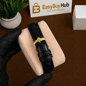 A black alligator leather watch strap with a gold buckle, held by a hand wearing a black glove over a beige cushion on a wooden desk. In the background, there is a small potted plant, a white box, and a sign that reads 'EasyBuyHub'.