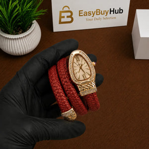 A red braided leather wrap watch with a gold-tone case and bezel, held in a hand wearing a black glove. The background shows a wooden table, a small potted plant, a branded EasyBuyHub card, and a white box.