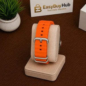 Bright orange silicone watch strap with a silver buckle, displayed on a beige watch stand on a dark wood table. A small potted plant is on the left, and a branded EasyBuyHub box is in the background.