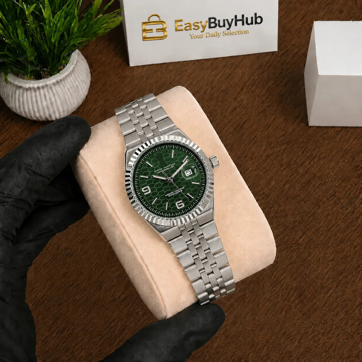 A silver stainless steel watch with a textured green dial and matching green bezel, held by a black-gloved hand on a beige cushion over a brown wooden surface. A white box with the 'EasyBuyHub' logo is visible in the background.