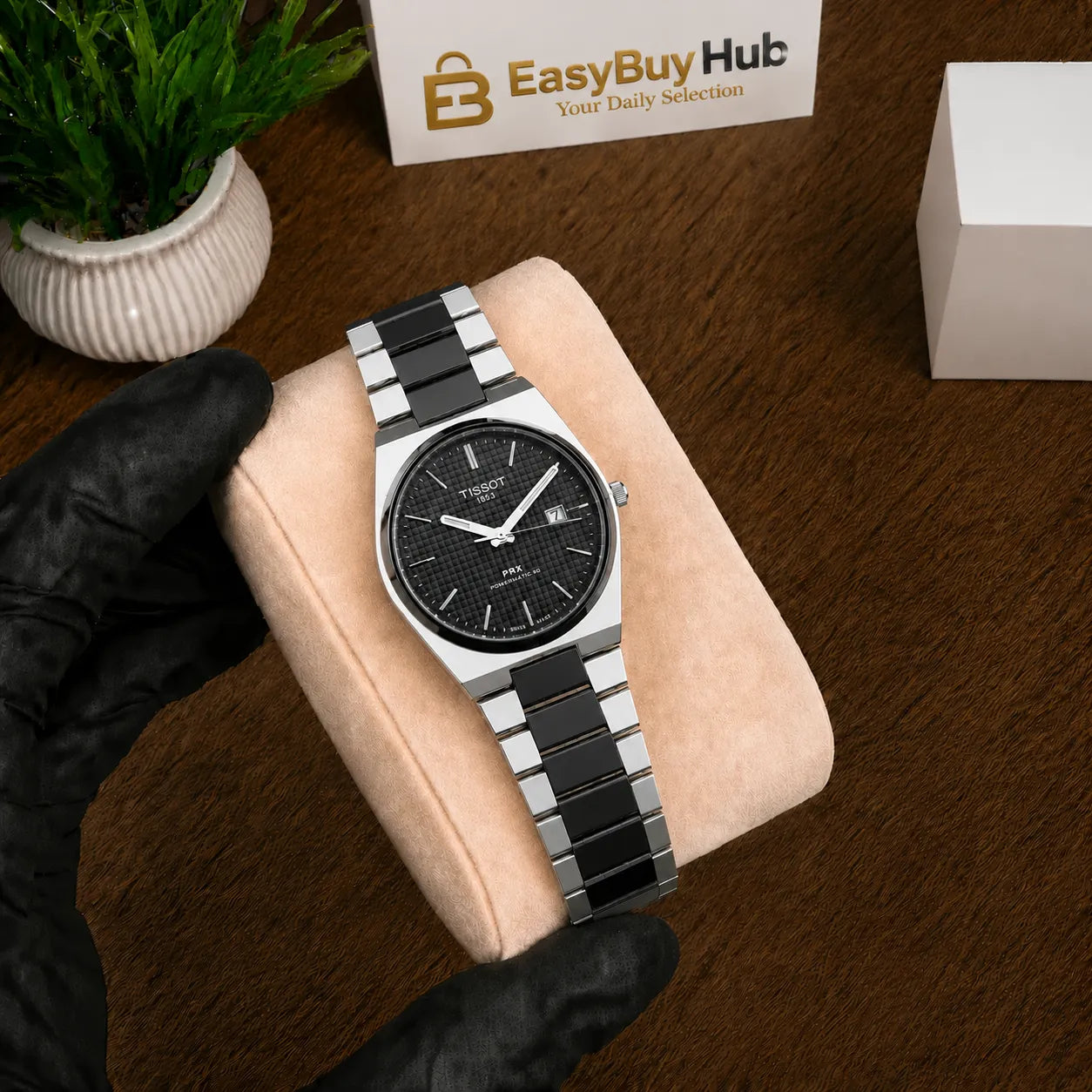 A stainless steel and black ceramic link watch with a black dial is displayed on a beige velvet watch cushion, held by a hand wearing a black glove. The background is a dark wood surface with a small potted plant and a branded 'EasyBuy Hub' box.