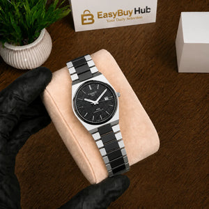 A stainless steel and black ceramic link watch with a black dial is displayed on a beige velvet watch cushion, held by a hand wearing a black glove. The background is a dark wood surface with a small potted plant and a branded 'EasyBuy Hub' box.