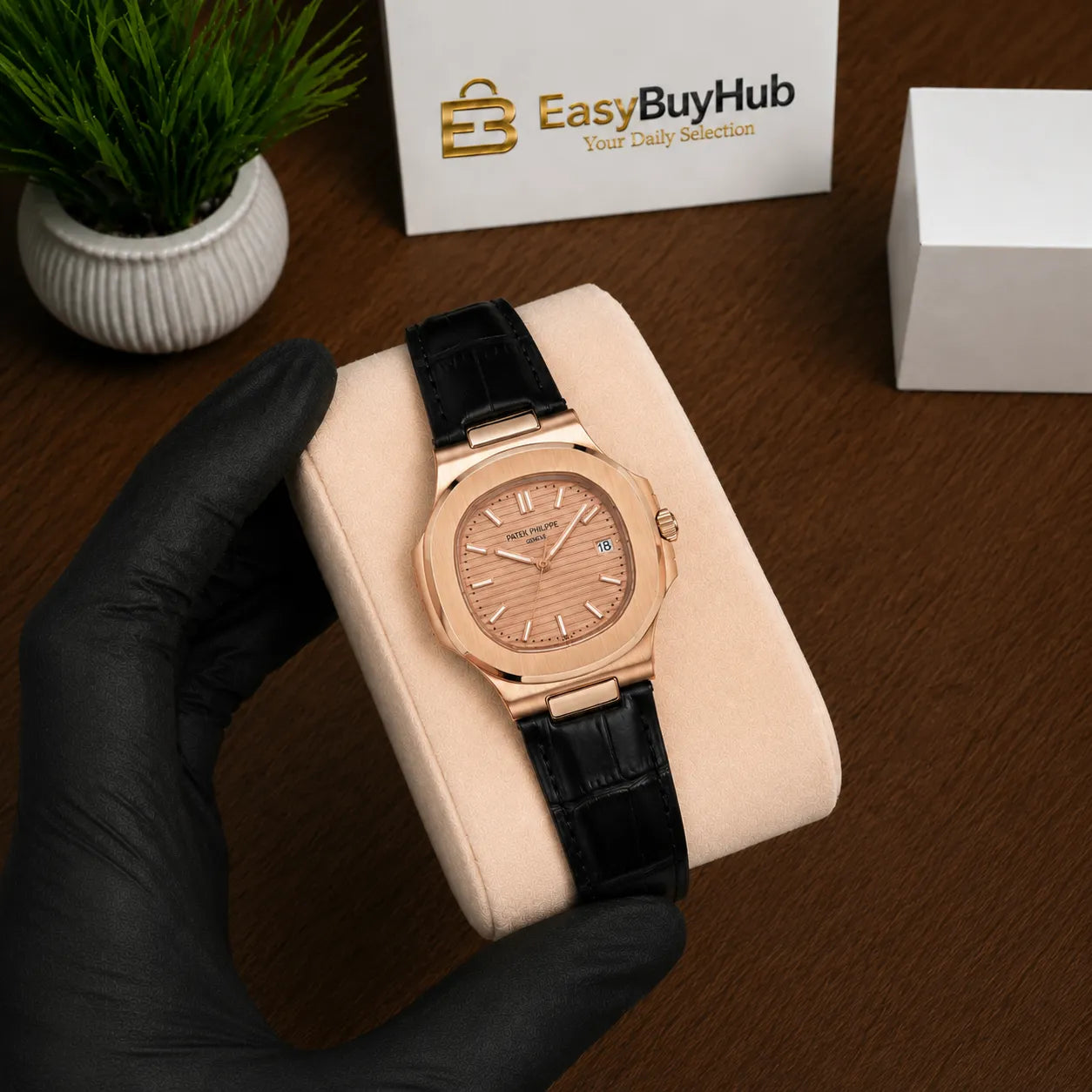 A rose gold watch with a black leather strap is displayed on a beige cushion held by a hand wearing a black glove. The background shows a wooden table, a small potted plant, a white box, and an EasyBuyHub card.