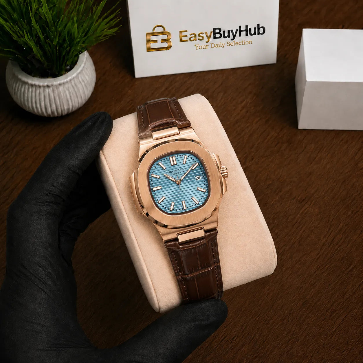 A rose gold watch with a light blue dial and brown leather strap, displayed on a cream cushion held by a hand wearing a black glove. A white box with the 'EasyBuyHub' logo is visible in the background on a wooden table.