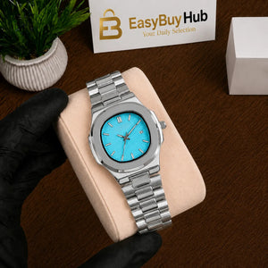 A silver stainless steel men's watch with a bright turquoise blue dial, resting on a beige display cushion held by a hand wearing a black glove. A wooden table surface, a small potted plant, and a branded 'EasyBuyHub' card are visible in the background.