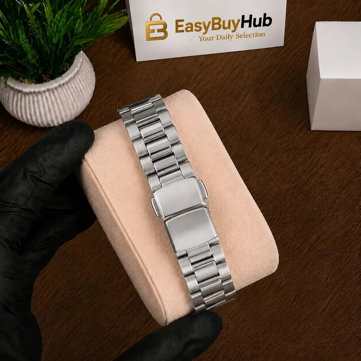 A silver stainless steel link watch band with a rectangular clasp, shown resting on a beige display cushion held by a hand in a black glove. A branded EasyBuyHub card and a small potted plant are visible in the background on a wooden surface.