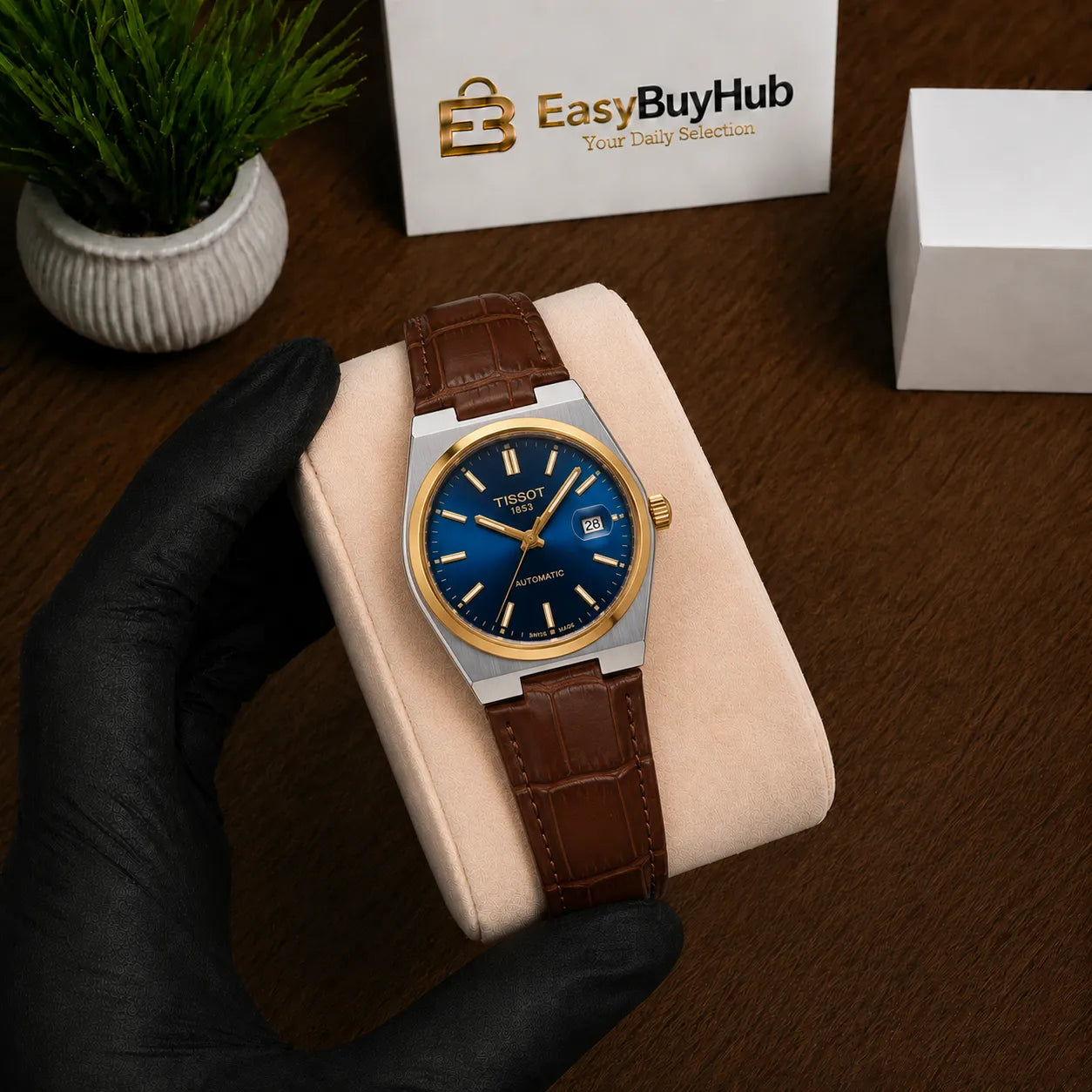 A Tissot watch with a blue dial, gold-tone accents, and a brown leather strap, displayed on a beige cushion held by a hand wearing a black glove. The background shows a wooden table, a small potted plant, and EasyBuyHub branding.