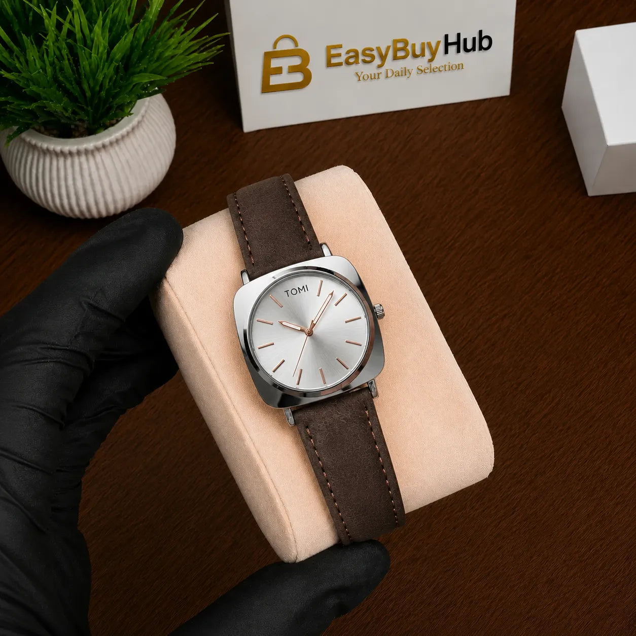 A silver-tone Tomhi analog watch with a brown leather strap and white dial, displayed on a beige cushion held by a hand in a black glove. A branded EasyBuyHub box and a small potted plant are visible in the background on a wooden surface.