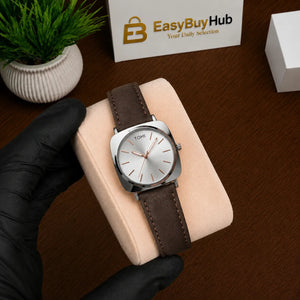 A silver-tone Tomhi analog watch with a brown leather strap and white dial, displayed on a beige cushion held by a hand in a black glove. A branded EasyBuyHub box and a small potted plant are visible in the background on a wooden surface.