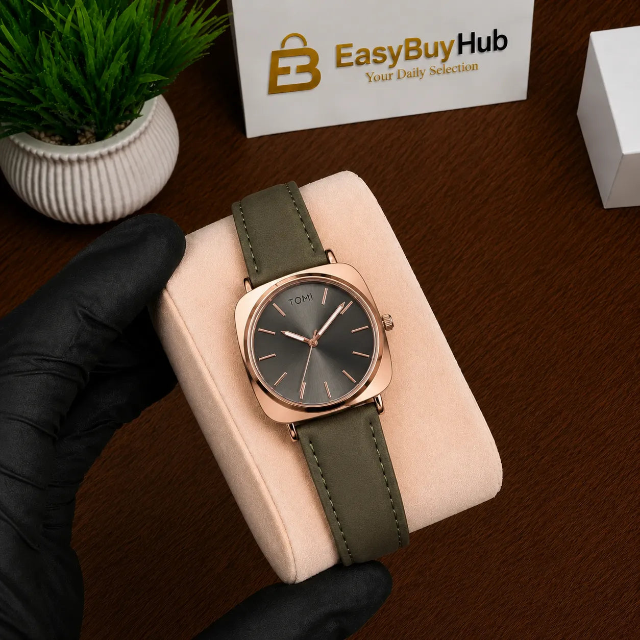 A wristwatch with a dark gray dial and olive green strap, held by a black-gloved hand on a beige display cushion over a brown wooden surface. A branded card reading EasyBuyHub is visible in the background.
