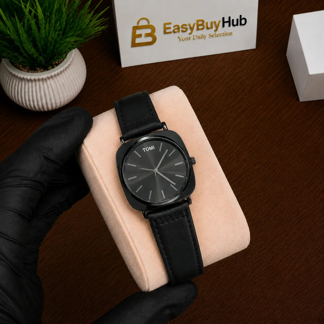 A black Tomsi watch with a black leather strap and a dark gray dial, resting on a light pink watch cushion held by a hand in a black glove. The background shows a dark wooden table, a small potted plant, and a box with the 'EasyBuyHub' logo.